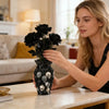 A woman holds a black decorative vase filled with dark flowers and skull decorations on a marble table.