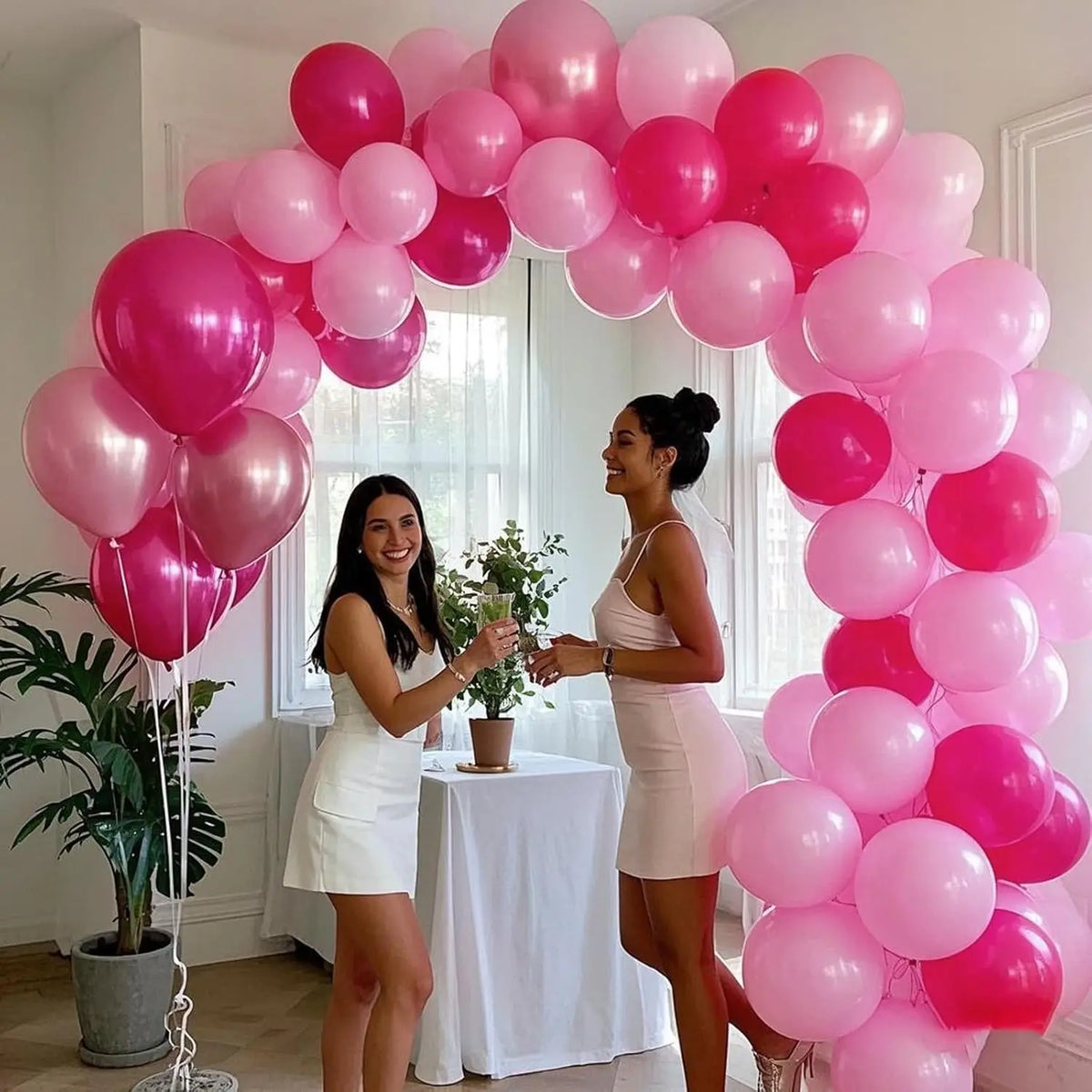 A woman in a white dress is holding flowers while another woman stands next to a large pink and magenta balloon garland.