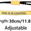 A gold-colored stainless steel bracelet with black cord and the text GOD IS IN CONTROL engraved on a bar.