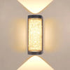 A vertical, cylindrical LED crystal outdoor wall sconce with a warm yellow glow mounted on a light beige wall.