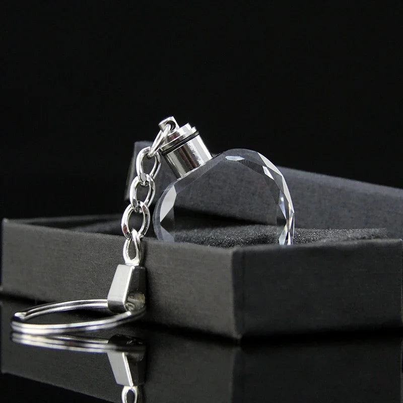 Llavero de cadena plateada con un dije de corazón de cristal transparente sobre una caja negra.