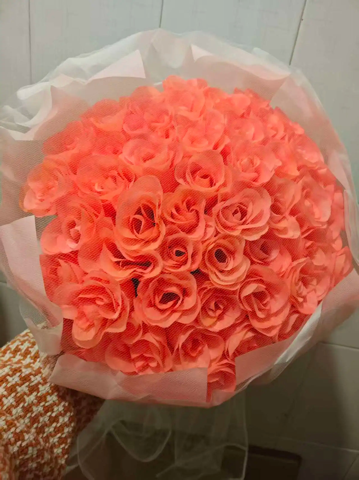 A bouquet of vibrant orange-pink silk roses wrapped in sheer white paper.