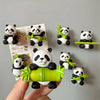 A hand holds a green panda magnet next to several other 3D panda fridge magnets on a gray wall.