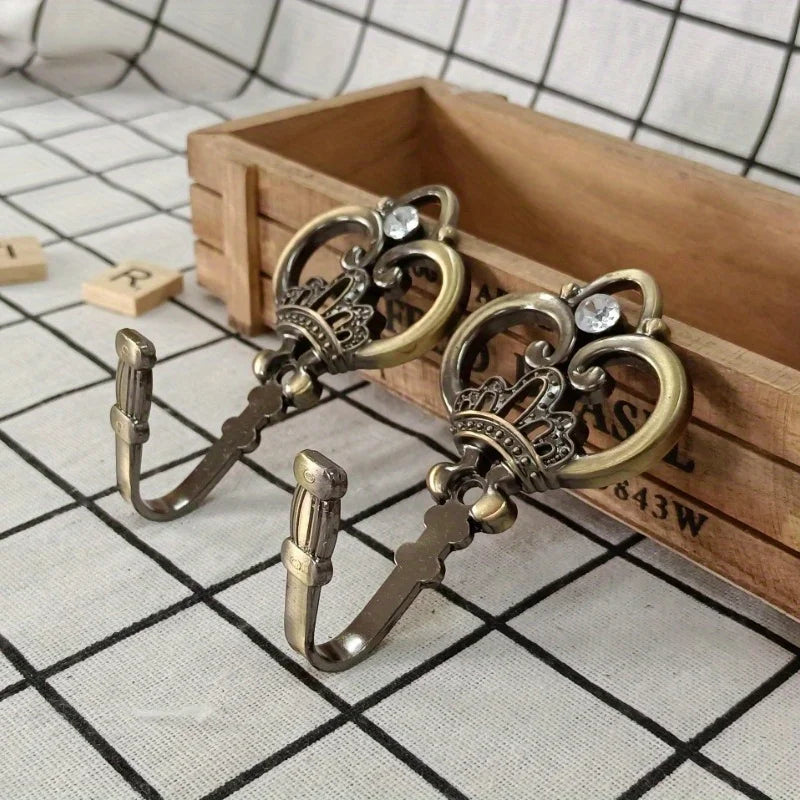 A pair of vintage crown curtain hooks with decorative metalwork resting in a wooden box on a checkered surface.