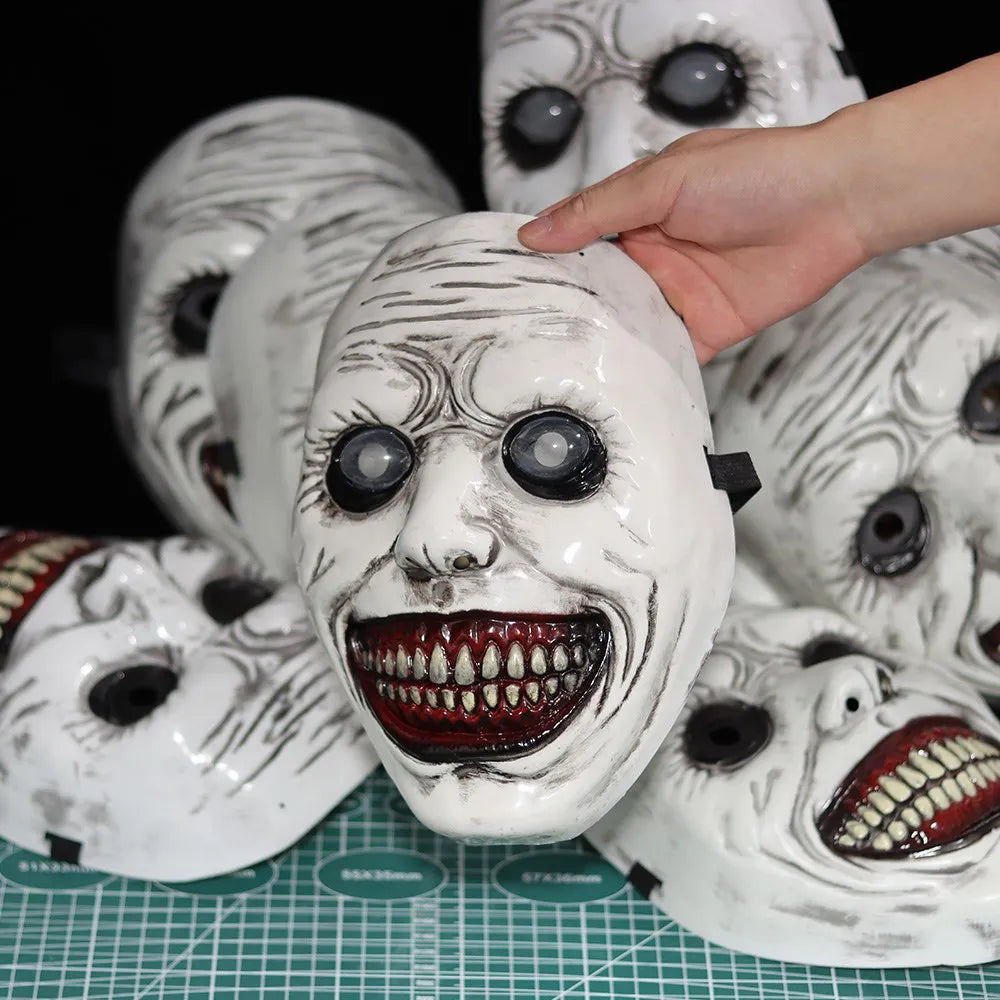 A white porcelain demon mask with facial markings and red teeth is being held by a hand against a dark background.