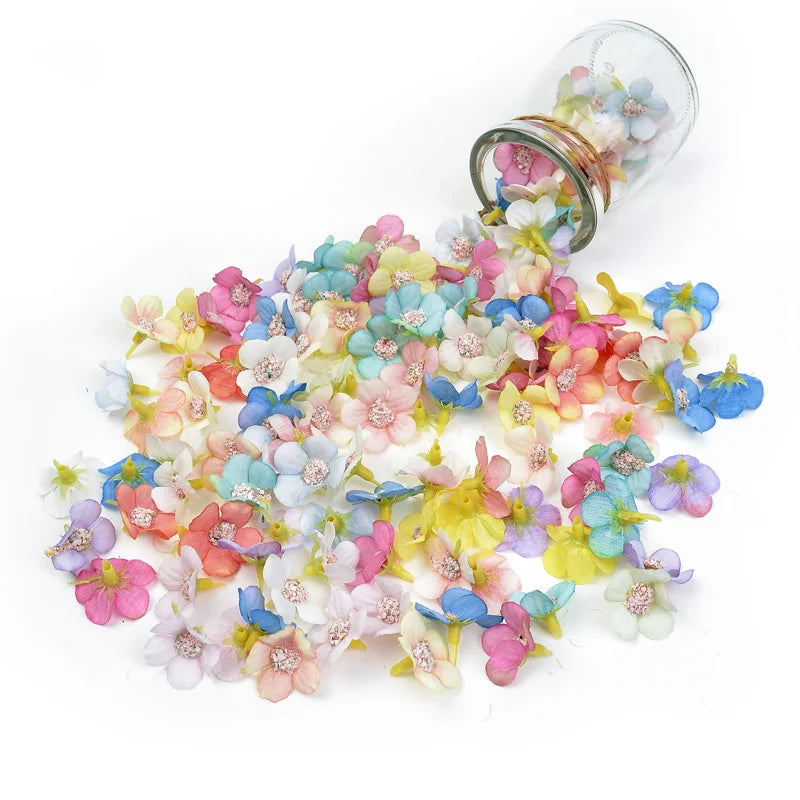 A pile of silk artificial flower heads in various pastel colors, including pink, blue, and yellow, next to a glass jar.