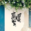 Black silhouette of an Art Nouveau butterfly design on a cream-colored wall, framed by green foliage.