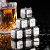 A stack of clear stainless steel ice cubes rests on a dark surface next to a glass of amber liquid.