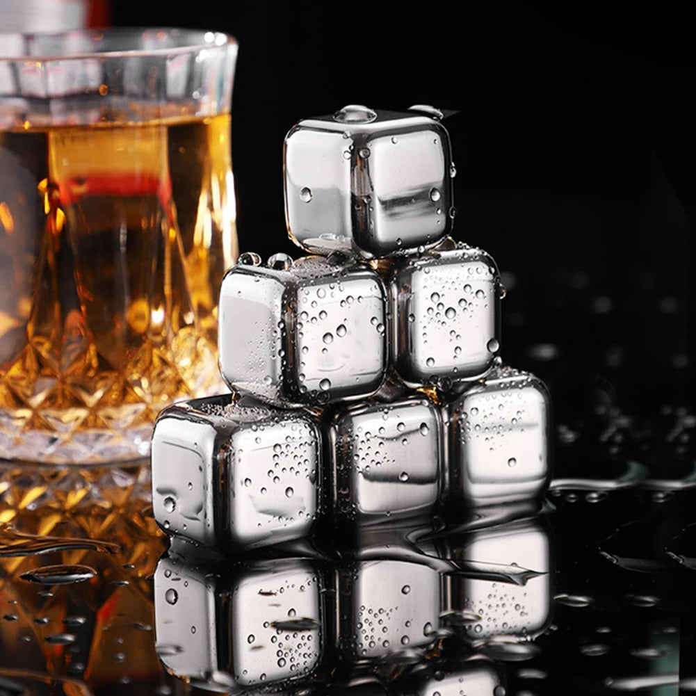 A stack of clear stainless steel ice cubes rests on a dark surface next to a glass of amber liquid.