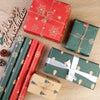 A collection of festive Christmas gift wrapping paper rolls and boxes in red, green, and gold tones on a wooden surface.