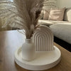 A tall dried floral arrangement in a white vase sits on a white ceramic stand next to a white ribbed concrete-style mold.