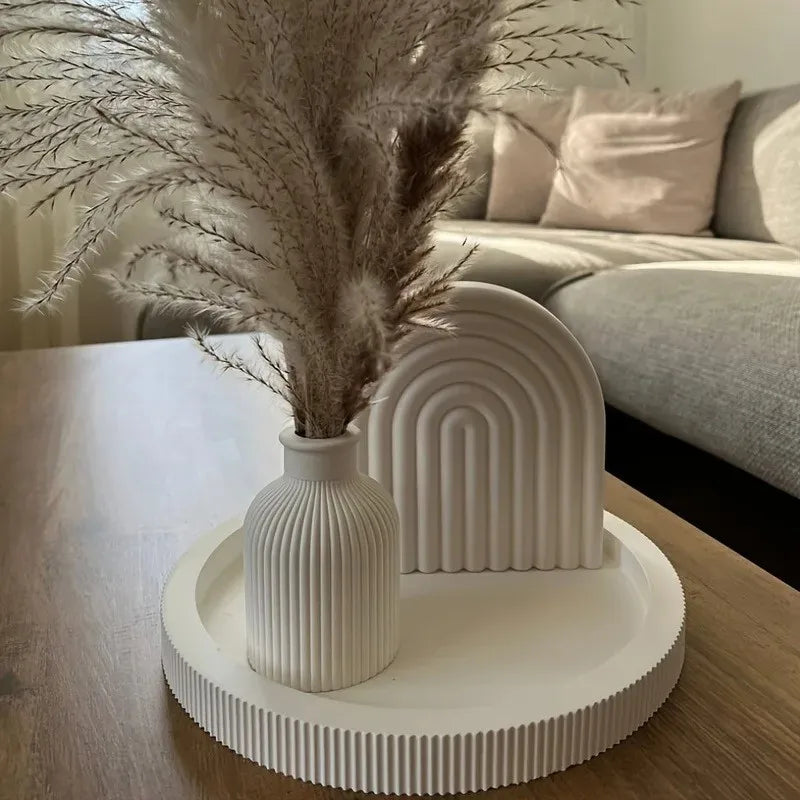 A tall dried floral arrangement in a white vase sits on a white ceramic stand next to a white ribbed concrete-style mold.