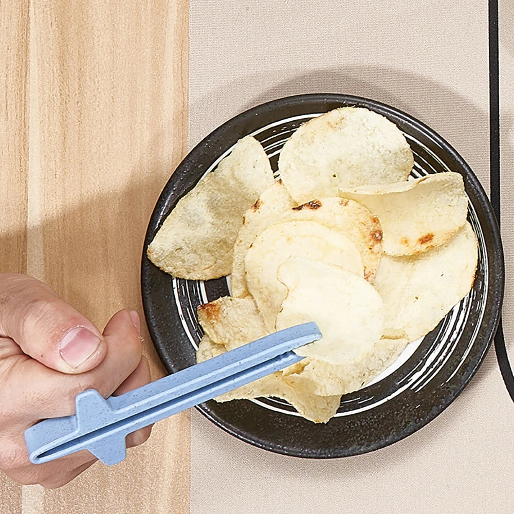A hand uses blue reusable wheat straw finger chopsticks to pick up potato chips in a black bowl.