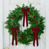A green evergreen wreath decorated with deep red velvet bows and pinecones, displayed on a white door.