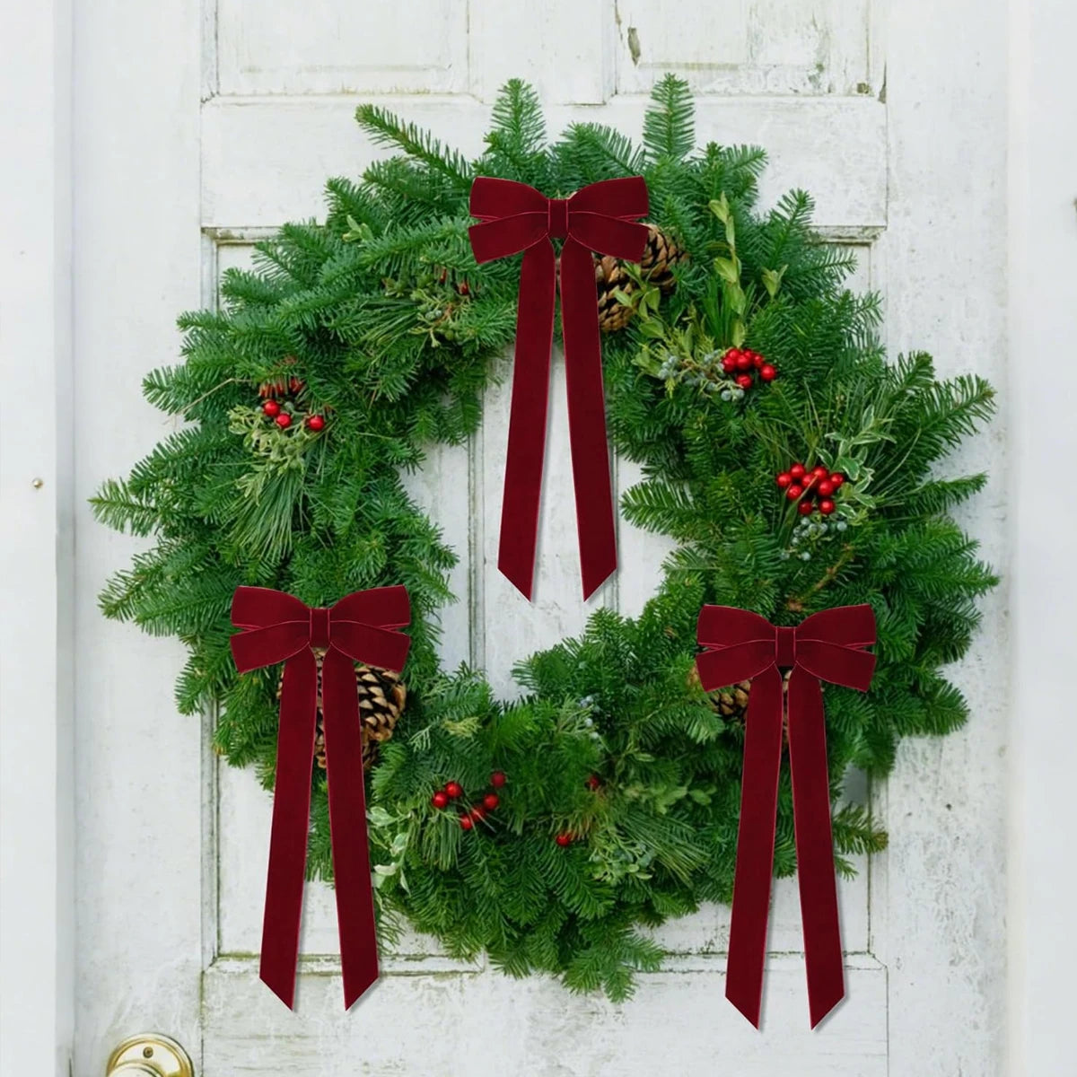 A green evergreen wreath decorated with deep red velvet bows and pinecones, displayed on a white door.