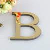 A 3D golden letter B cutout for a mirror wall decal set, resting on a white surface with flowers in the background.