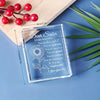 A clear crystal keepsake with engraved text for a sister, resting on a wooden board next to green leaves.