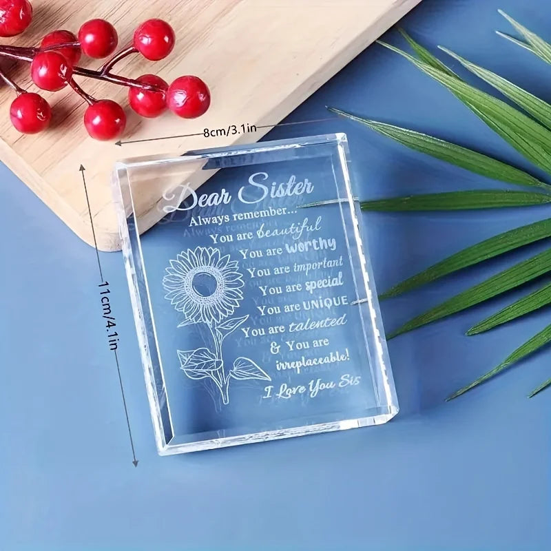 A clear crystal keepsake with engraved text for a sister, resting on a wooden board next to green leaves.