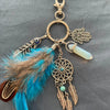 A bohemian dream catcher keychain featuring a feather, tassel, and light green bead on a gold-toned metal chain.