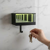 A hand is holding a gold-colored keychain with a loop, positioned near a black wall-mounted light fixture featuring an illumi
