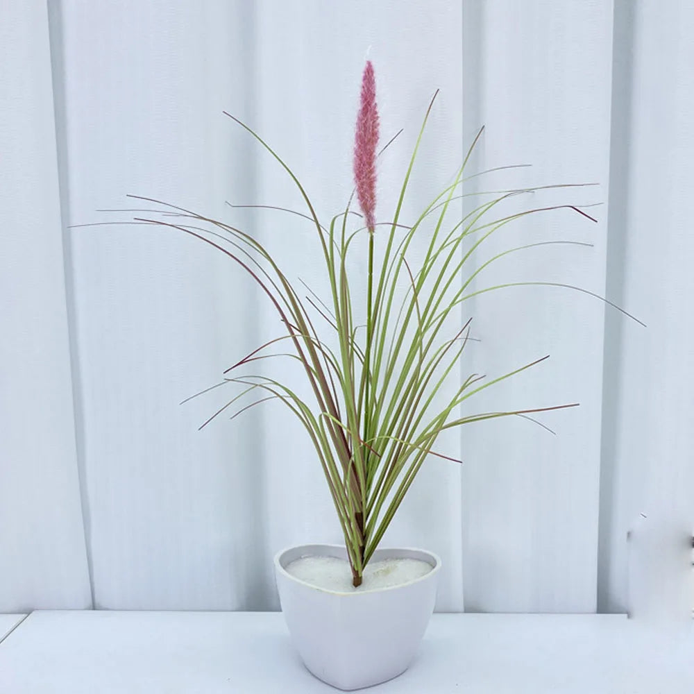 A potted artificial grass plant with long green blades and a tall pink flower spike on a white surface.