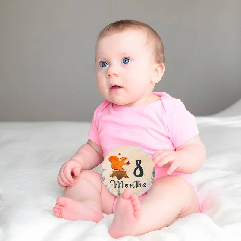 A baby sitting on a white surface looking to the side next to a wooden milestone card showing 8 months.