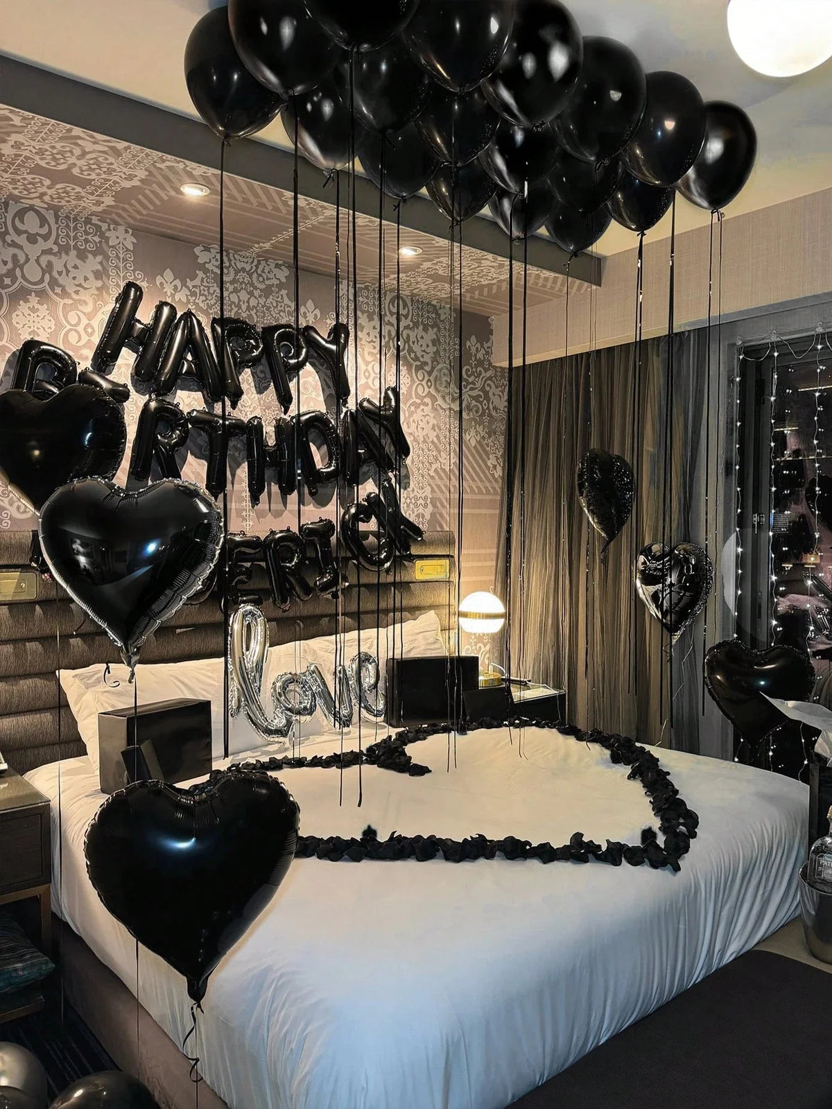 A bedroom decorated with black balloons spelling out Happy Birthday and love, featuring a bed with white linens.