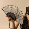 A hand holding an antique style bamboo folding fan with intricate black and white line art designs.