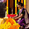 A woman in a purple and green saree is arranging bright orange and yellow artificial carnation and marigold flower heads for 