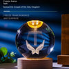 A glass crystal ball night light with a glowing golden top and a silver cross and wings design on a wooden base.