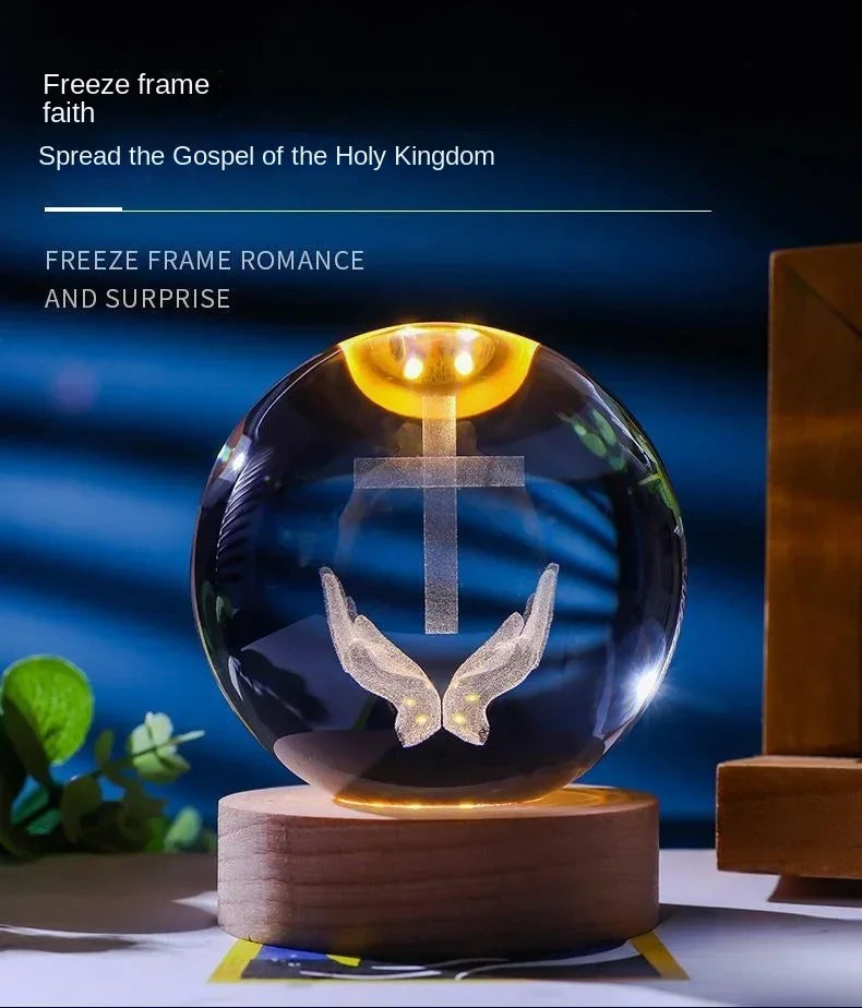 A glass crystal ball night light with a glowing golden top and a silver cross and wings design on a wooden base.