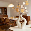 A white, modern heart-shaped vase holds dry, golden-brown branches and leaves on a wooden table.