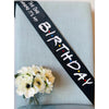 A black ribbon with white text reads Birthday where it's my on a light blue fabric background with white flowers.