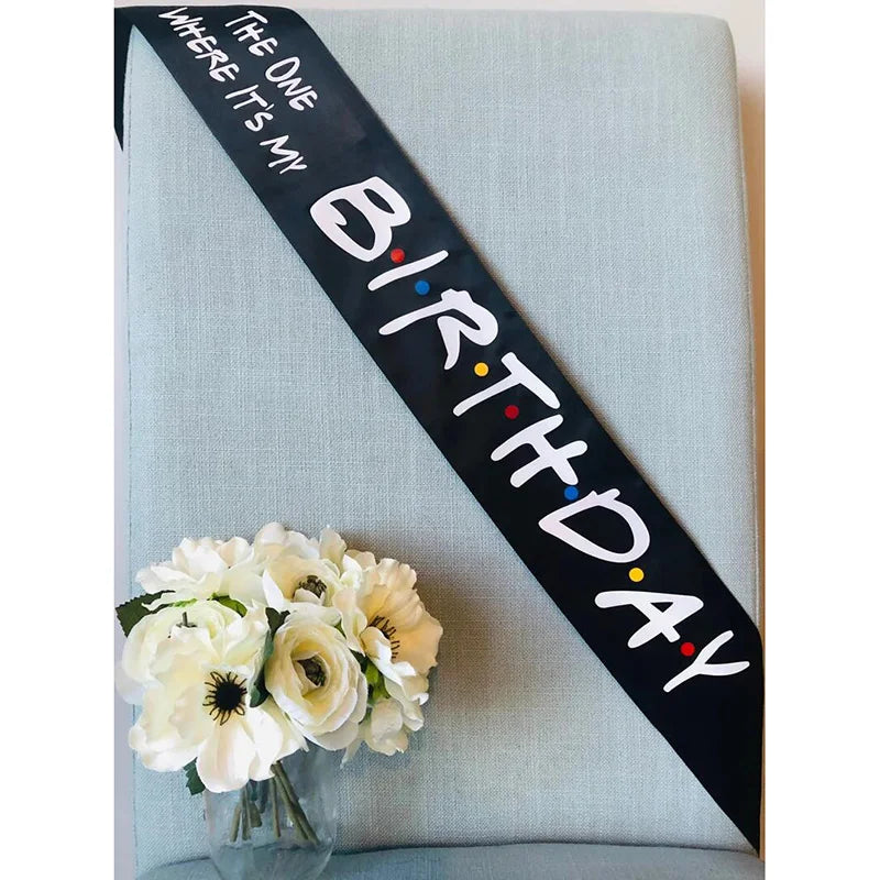 A black ribbon with white text reads Birthday where it's my on a light blue fabric background with white flowers.