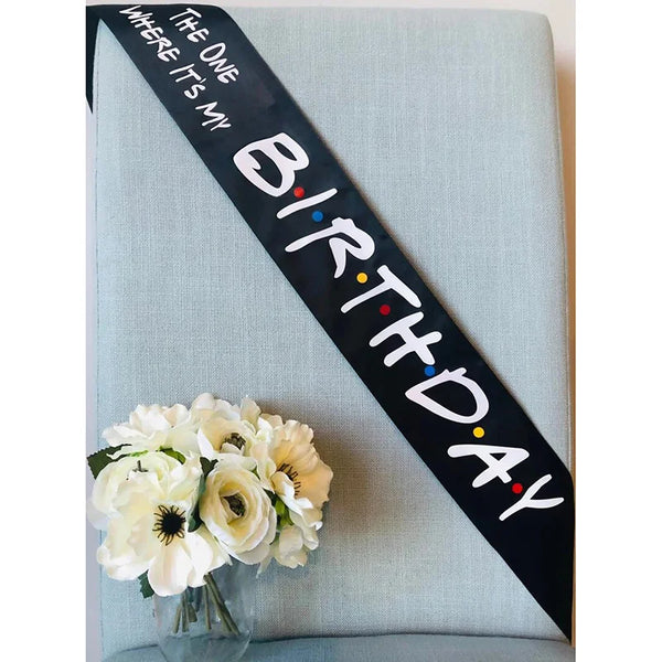 A black ribbon with white text reads Birthday where it's my on a light blue fabric background with white flowers.