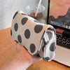 A hand holds a beige canvas travel makeup bag with black polka dots, next to a laptop.