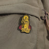 A gold enamel pin depicting Saint Judas Tadeo with a halo and olive branch on a fabric background.