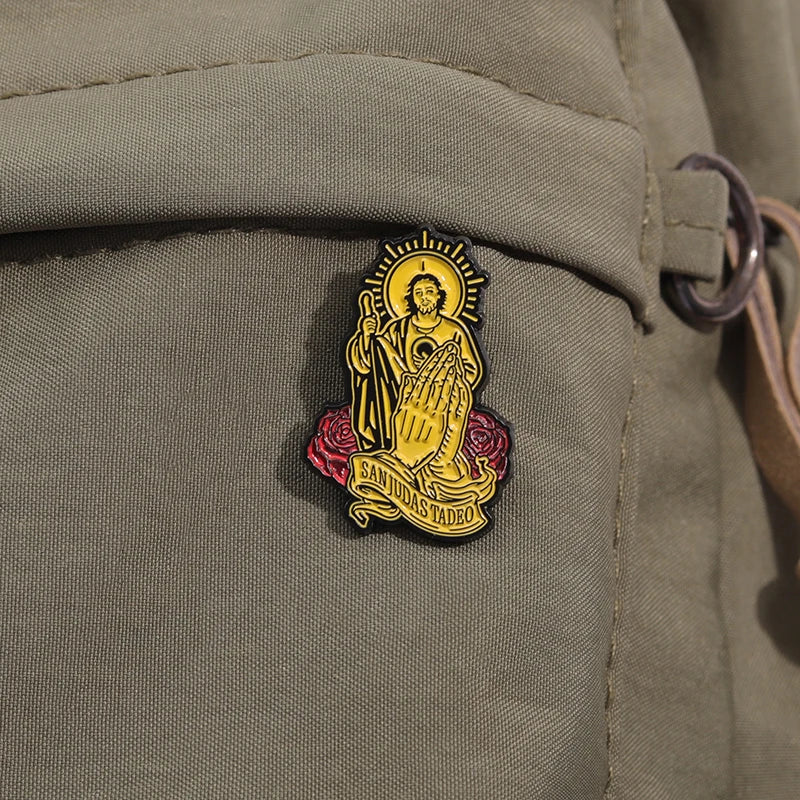 A gold enamel pin depicting Saint Judas Tadeo with a halo and olive branch on a fabric background.