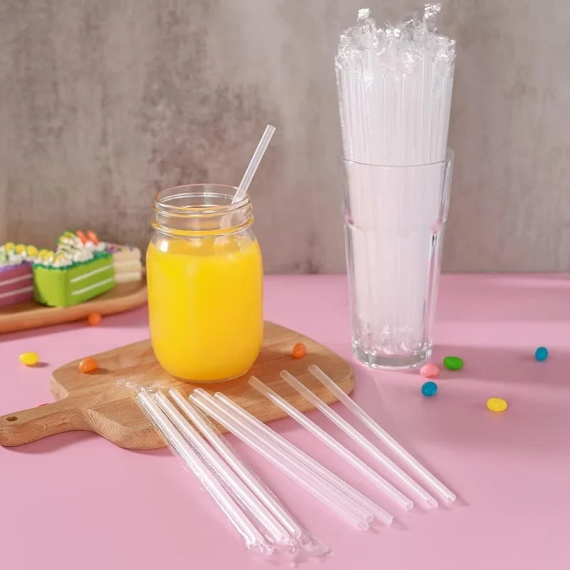 A glass jar of orange juice next to a clear glass filled with ice and plastic drinking straws on a pink surface.