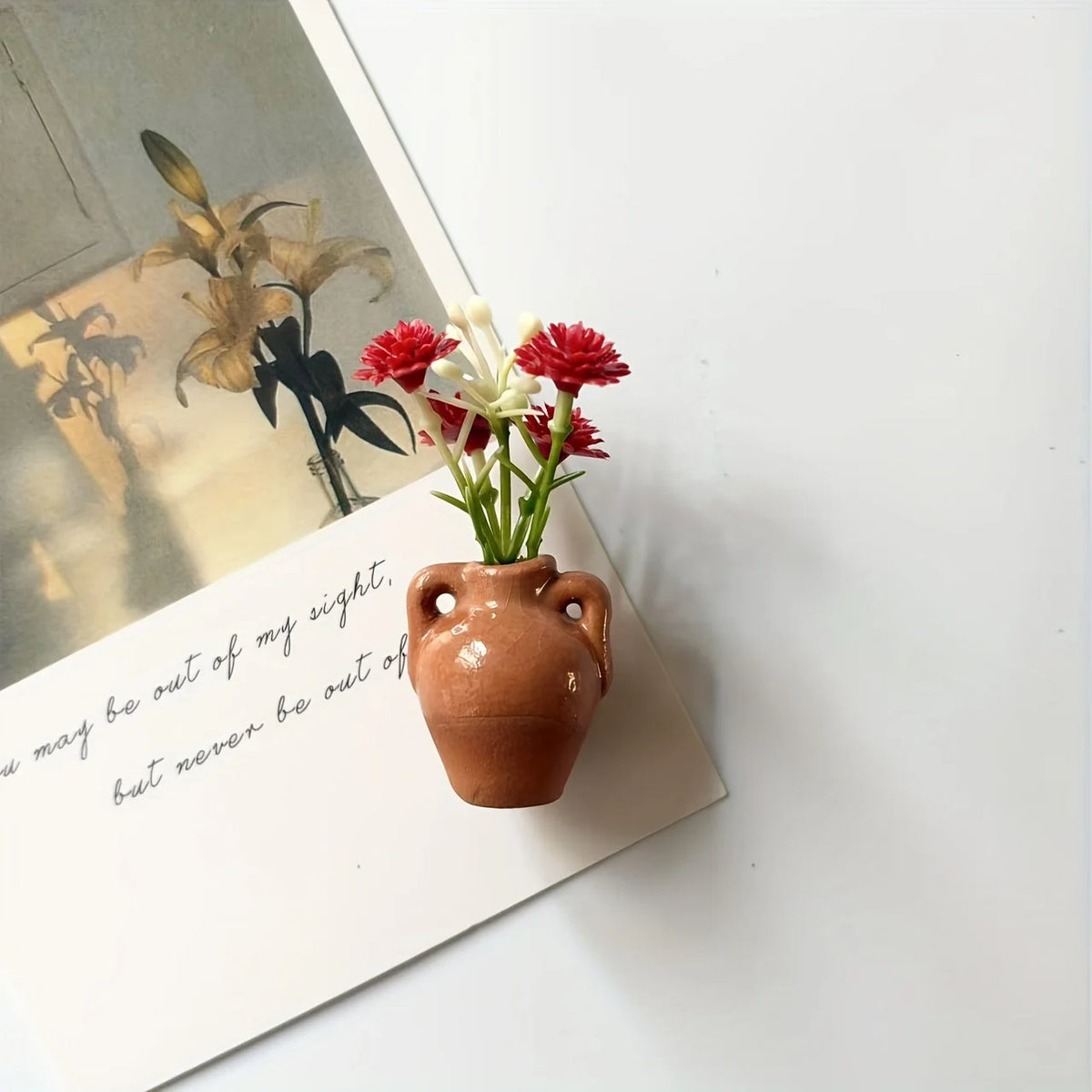 A small terracotta ceramic vase holding red and white artificial flowers, placed on a white card.