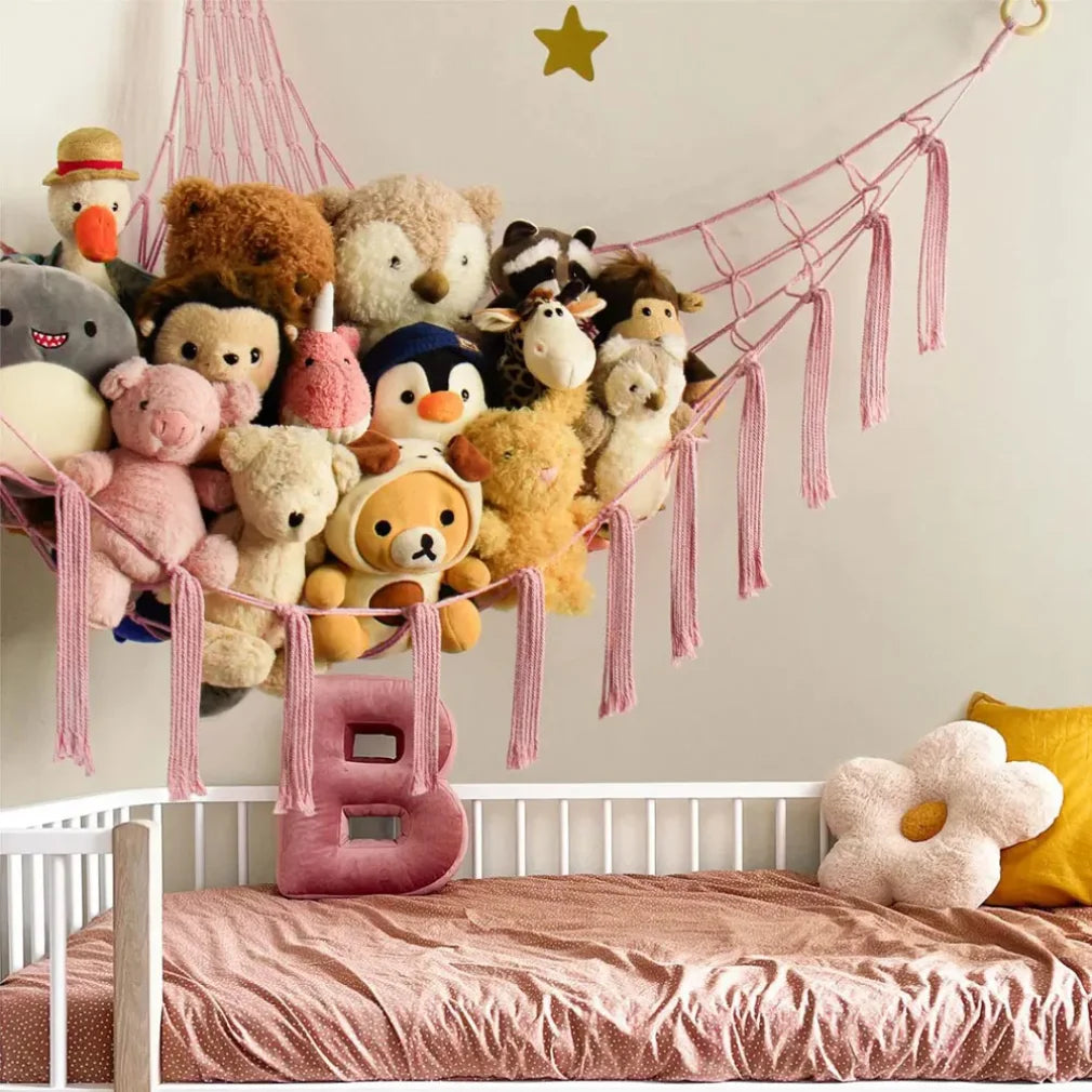 A multi-use storage hammock made of pink fabric is suspended above a bed, filled with various stuffed animals and plush toys.