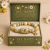 A gold stretch charm bracelet with green beads and a bee charm displayed in a natural green box.