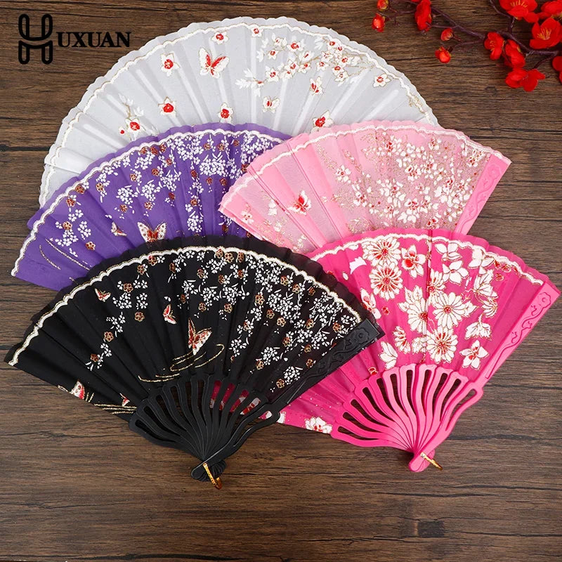 A collection of five elegant plum blossom folding fans in white, purple, pink, and black on a wooden surface.