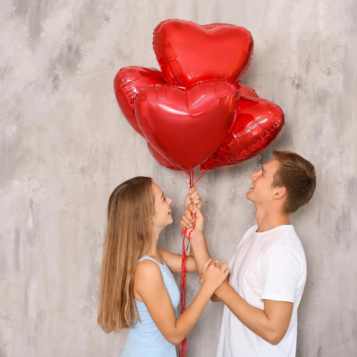 A young couple is holding together a cluster of red heart foil balloons against a gray background.