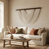 A bohemian macrame wall tapestry hangs on a beige wall above a light sofa and wooden coffee table.