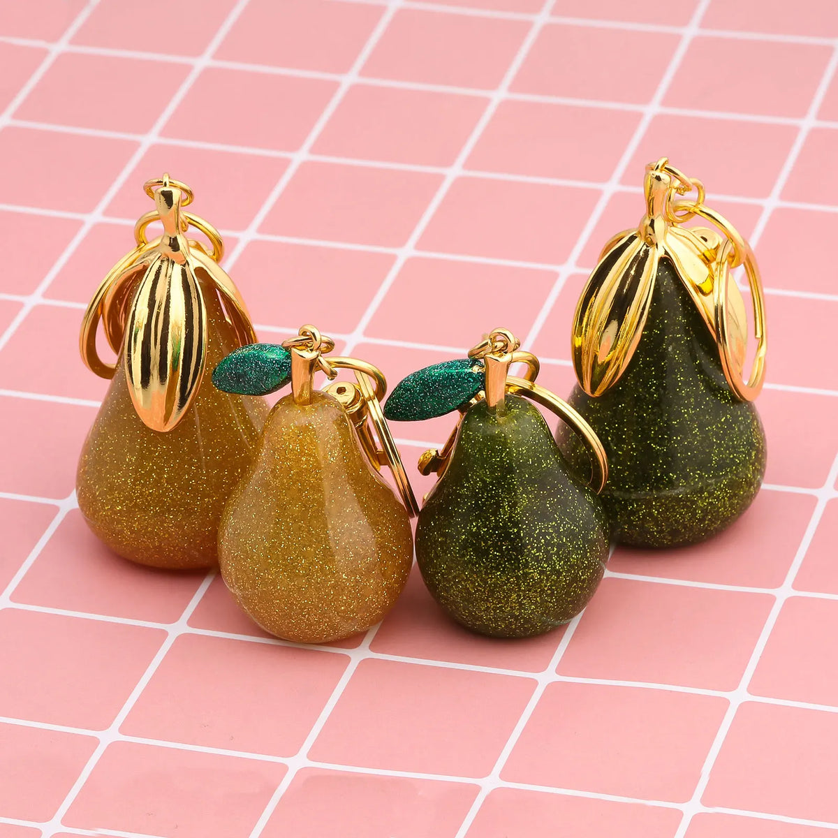 Three pear-shaped fruit keychains with gold findings and glittery green and gold finishes on a pink grid background
