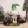 A festive green and white gnome decoration stands next to a vintage red wagon in a snowy outdoor setting.