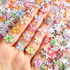 Una mano muestra 50 adornos de uñas de tortuga mini coloridos con diseños florales sobre un fondo texturizado.