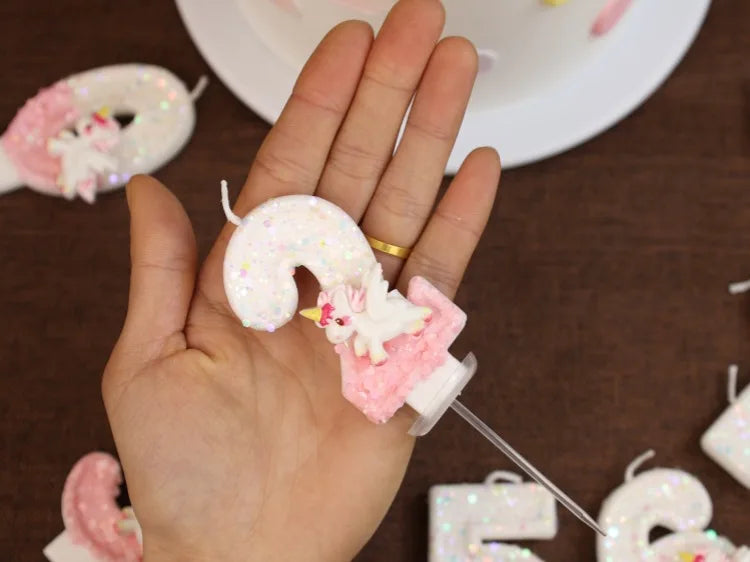 Una mano sostiene una vela mágica rosa y blanca decorada con purpurina y una pequeña figura.