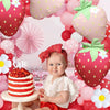 A baby is sitting in front of a large pink strawberry foil balloon surrounded by various pink and red balloons for a themed p